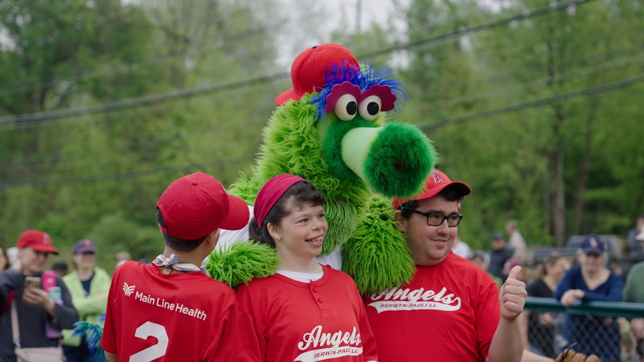 Three people in red Angels shirts pose with a bright green furry dragon mascot at an outdoor community event in a park.